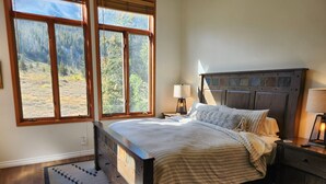 Une chambre à coucher avec un lit en bois, une table de chevet, une lampe et une vue sur une forêt, visible à travers de grandes fenêtres.