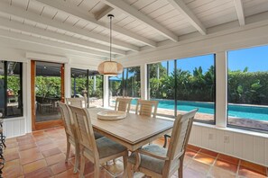 Un espace repas comprenant une grande table et des chaises, un luminaire suspendu, et une vue sur une piscine et la verdure environnante.