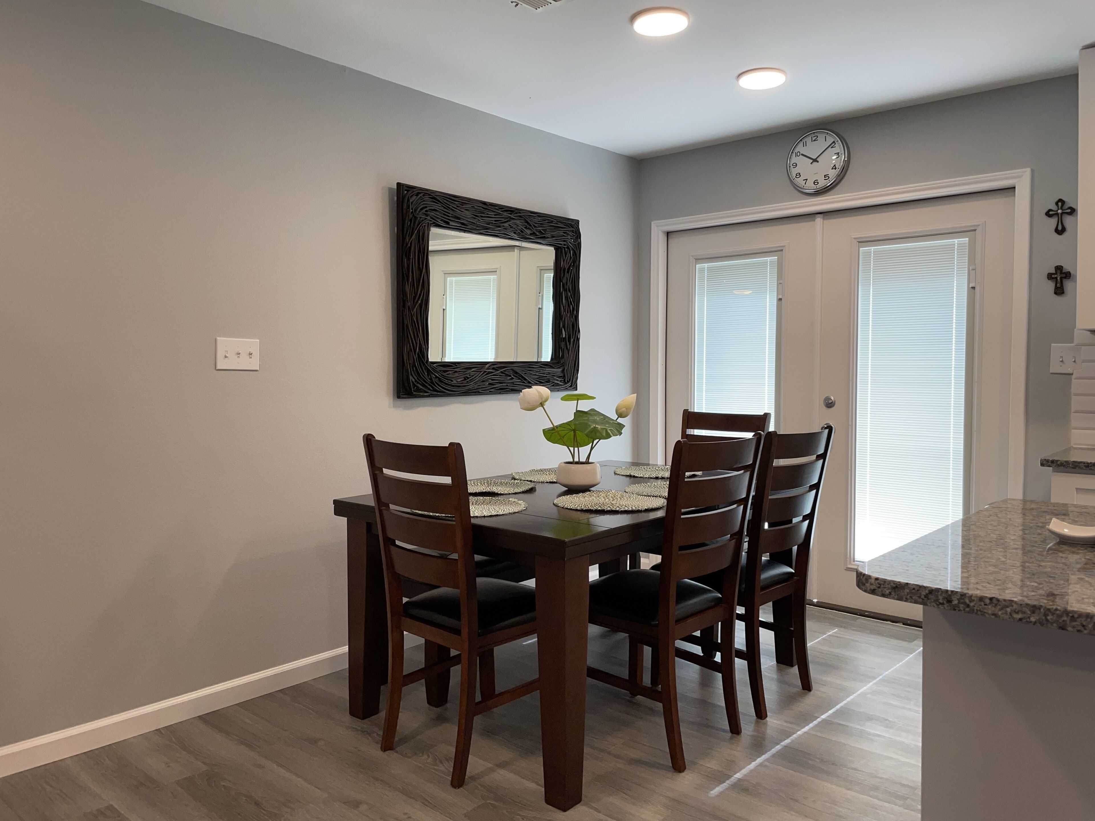 A dining area with a dark wooden table and chairs, a mirror on the wall, and a clock above the door.