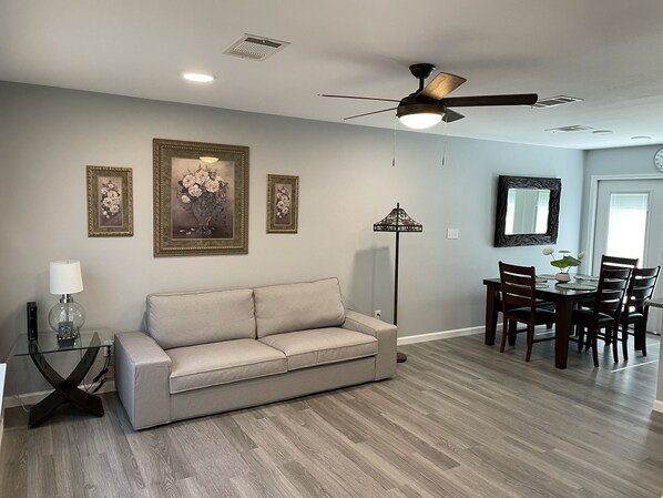 A living room with a beige sofa, a dining table with chairs, a ceiling fan, and framed floral artwork on the wall.