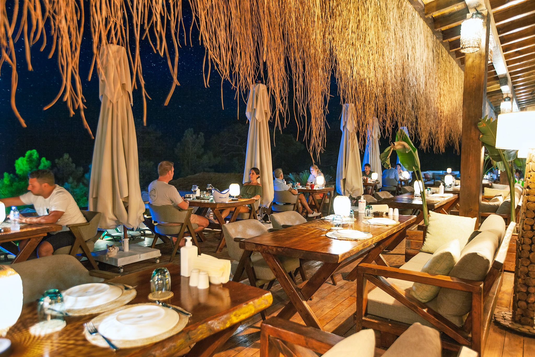An outdoor dining area with wooden tables and chairs, covered by thatched umbrellas, and a view of the night sky.