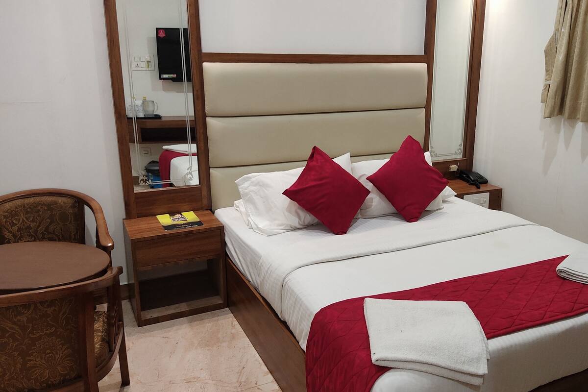 A hotel room with a bed, a wooden nightstand, a chair, a desk, a mirror, and a television.