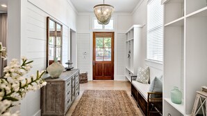 A hallway with white walls, a wooden door, a chandelier, a wicker bench, and a large woven rug. - Stunning WC Home  | Private 6-Seat Golf Cart + Adult Bikes | Steps from Camp (Santa Rosa Beach)