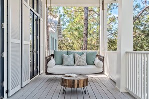 A porch with a swing seat, a round wooden coffee table, and a view of trees and a building in the background.