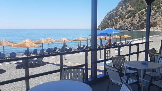 Um restaurante à beira-mar com mesas ao ar livre, guarda-sóis e vista para o mar e falésias rochosas.