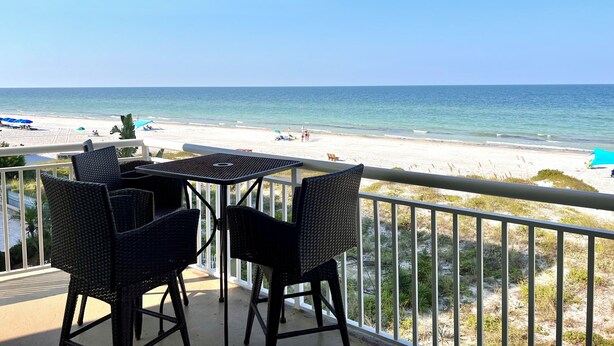A balcony with wicker chairs and a table overlooking a beach with umbrellas and people.