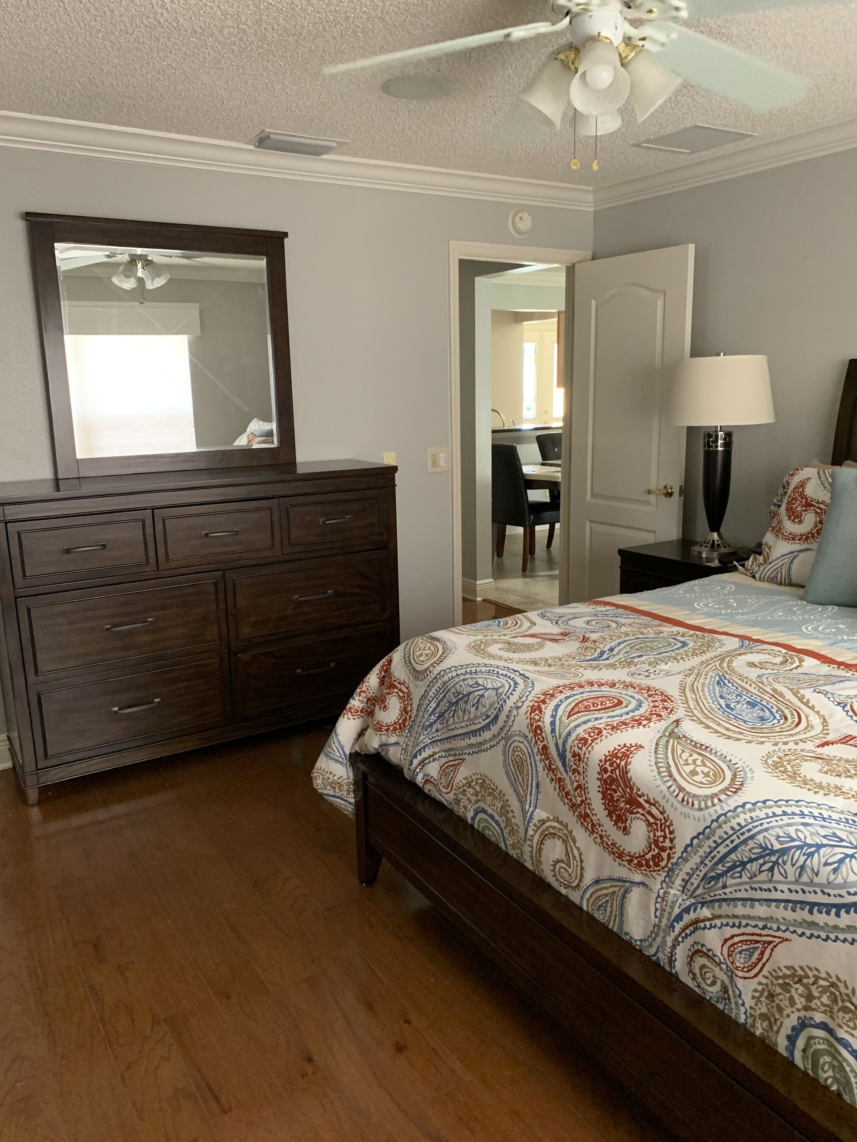A bedroom with a wooden dresser, a bed with a patterned bedspread, a ceiling fan, and a nightstand with a lamp.