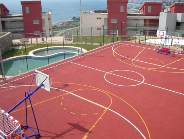 Uma quadra de basquete ao ar livre, com piscina e vista para prédios ao fundo.