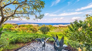 A tropical garden with a peacock sculpture, lush greenery, and a view of the ocean and distant hills. - Now Renting! Kapalua Ridge 2BR/3BA Villa Ocean Views, Pools & Beach Access (Lahaina)