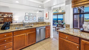 A modern kitchen with granite countertops, stainless steel appliances, and wooden cabinetry. There is a dining area with a view of the ocean. - Now Renting! Kapalua Ridge 2BR/3BA Villa Ocean Views, Pools & Beach Access (Lahaina)