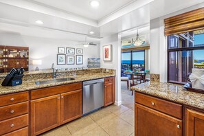 Cozinha moderna com bancadas de granito, eletrodomésticos de aço inoxidável e armários de madeira. Há uma área de jantar com vista para o oceano.