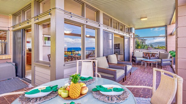 A covered outdoor patio with wicker furniture, a dining table set with fruit, and a view of the ocean. - Now Renting! Kapalua Ridge 2BR/3BA Villa Ocean Views, Pools & Beach Access (Lahaina)