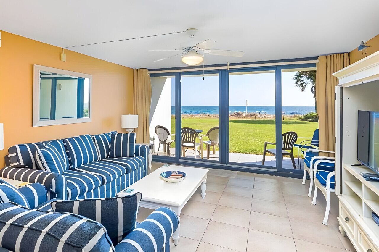 A room with a large sofa, a coffee table, and a TV. There is a large window with a view of the ocean and a grassy area.
