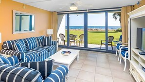 A room with a large sofa, a coffee table, and a TV. There is a large window with a view of the ocean and a grassy area. - The best location in Orange Beach - Ground floor condo - NEW to rental market (Orange Beach)