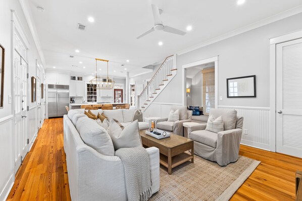 A spacious living room with a sofa, armchairs, and a coffee table. A staircase leads to the upper floor, and a kitchen is visible in the background.