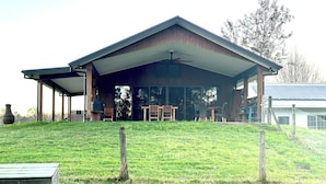 A covered outdoor area with a table and chairs, a barbecue grill, and a large window overlooking a grassy field. - Riverfront Farm Stay with Animal Nursery at The Hatch Farm, Port Macquarie (Blackmans Point)