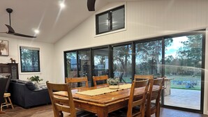 A dining area with a wooden table and chairs, a sofa, and a view of a backyard through large windows. - Riverfront Farm Stay with Animal Nursery at The Hatch Farm, Port Macquarie (Blackmans Point)