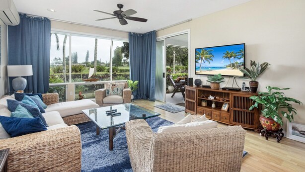 Amplia sala de estar con una gran mesa de centro de vidrio, muebles de mimbre, ventilador de techo y vistas al exterior.
