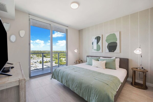 A bedroom with a large bed, a desk with a computer, a TV, and a view of a cityscape through a sliding door.