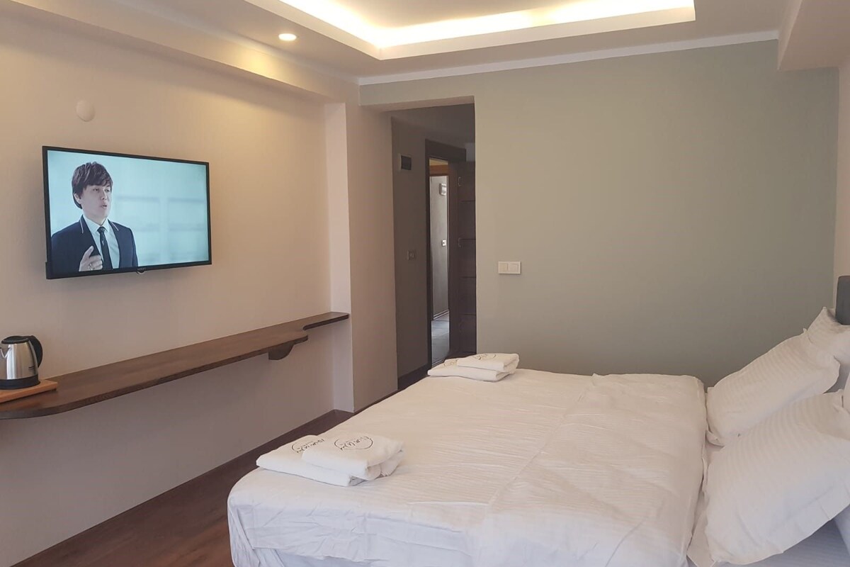 Habitación de hotel con una cama, un televisor en la pared, una cafetera y una puerta que da a otra habitación.