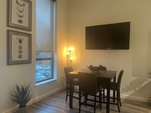 A dining area with a table and chairs, a potted plant, framed artwork, a window with a view of parked cars, and a wall-mounted flat-screen TV.