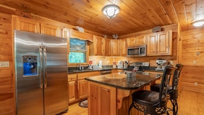 A wooden cabin kitchen with a stainless steel refrigerator, wooden cabinets, a granite countertop, and a bar stool. - Riverfront • Pets • Fire Pit • Free Night (Ellijay)
