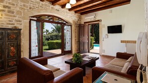 A spacious living room with a stone wall, wooden ceiling beams, a large window, and a view of a pool.