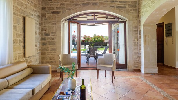 A stone-walled room with a sofa, two chairs, a table with a vase and a bottle, and a view through an arched doorway to an outdoor area with a table and chairs.