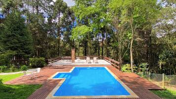 A rectangular swimming pool with a wooden deck surrounding it, set in a lush green garden with trees and a clear blue sky.