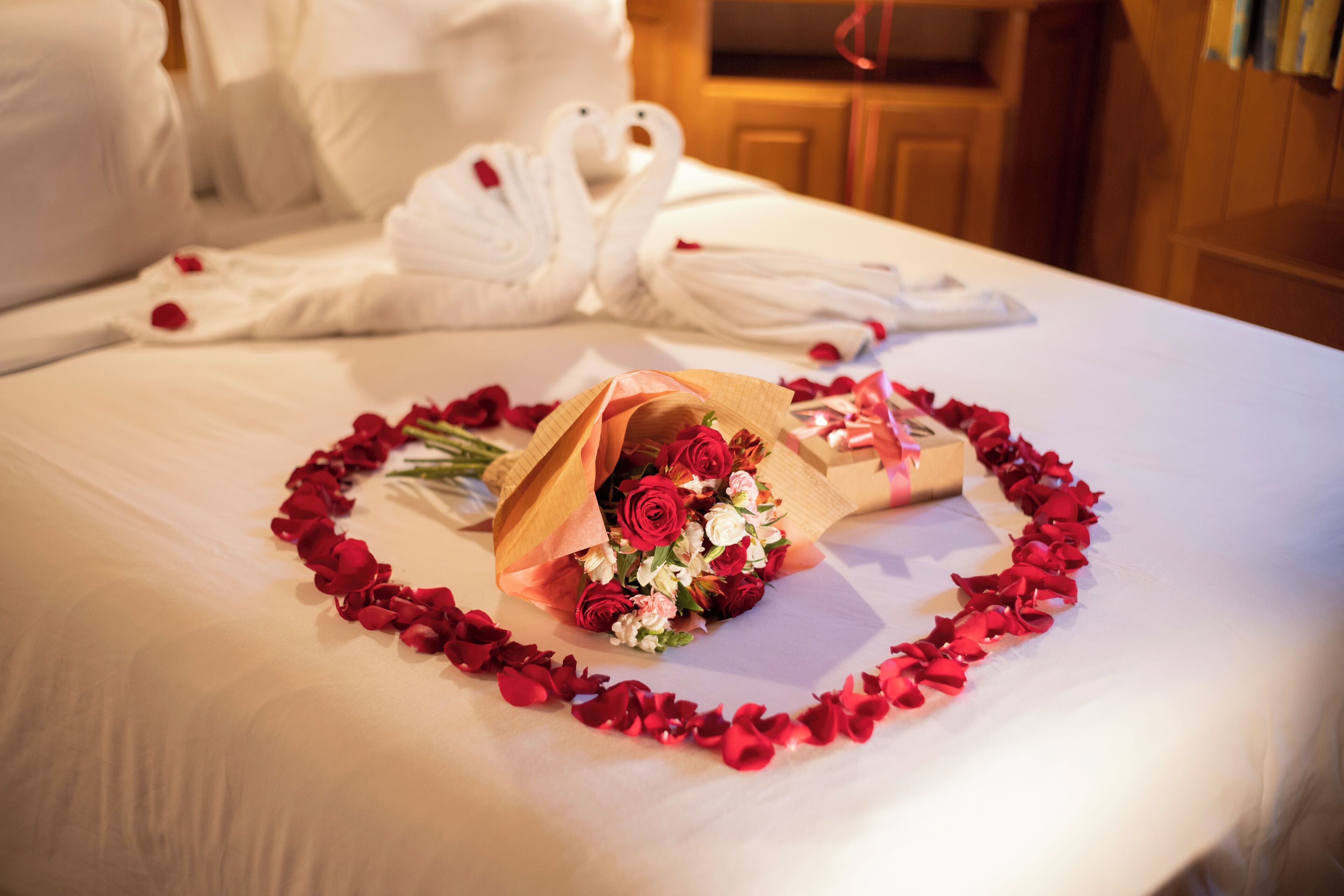 A bed with rose petals arranged in a heart shape, a bouquet of flowers, and a pair of white bathrobes.