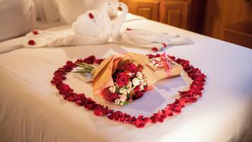 A bed with rose petals arranged in a heart shape, a bouquet of flowers, and a pair of white bathrobes.