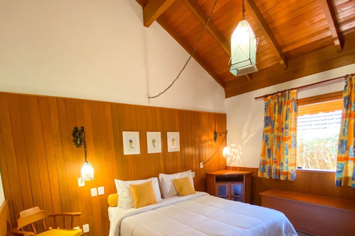 Un dormitorio con techo de madera, una cama con ropa de cama blanca, una silla de madera y una mesita de noche con una lámpara.