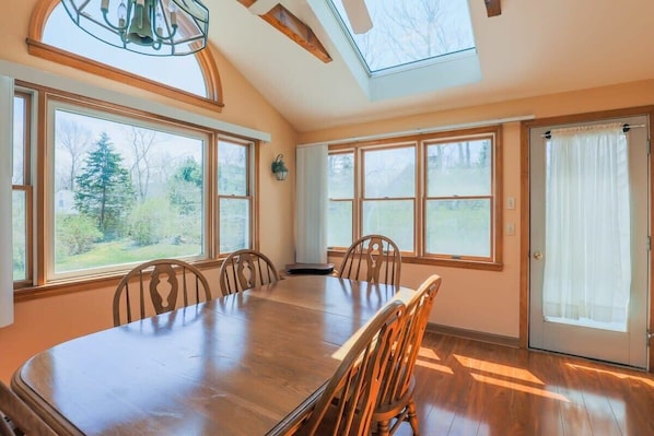 A dining room with a large wooden table and chairs, large windows with a view of trees, and a skylight.