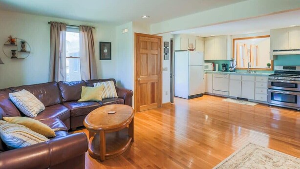 A living room with a brown leather sofa, a round wooden coffee table, and a kitchen with white cabinets and a stove. - Book Eastham, Plenty Of Room For Entire Family! (Eastham)