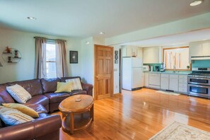 A living room with a brown leather sofa, a wooden coffee table, and a kitchen area with white cabinets and stainless steel appliances.