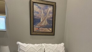 Ein ordentlich bezogenes Bett mit einer weißen Decke, ein gerahmtes Bild eines Segelboots an der Wand und eine Lampe auf einem Nachttisch.