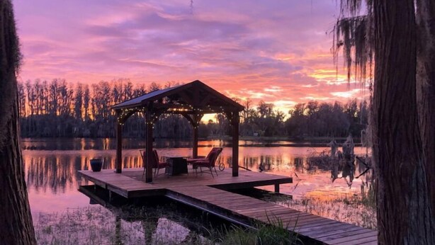 Eine Holzterrasse mit einem Pavillon, Stühlen und einem Tisch am Ufer eines ruhigen Sees bei Sonnenuntergang.