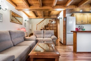 A cozy living room with a wooden ceiling, a glass-top coffee table, and a sofa. A staircase leads to a kitchen with wooden cabinets and a refrigerator.