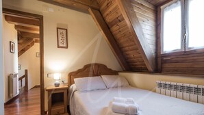 A bedroom with a wooden ceiling, a bed with white bedding, a nightstand with a lamp, and a window with lace curtains.