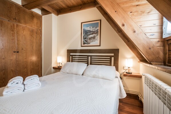 Une chambre avec un plafond en bois, un lit avec une literie blanche, deux lampes de chevet et un tableau encadré au mur.