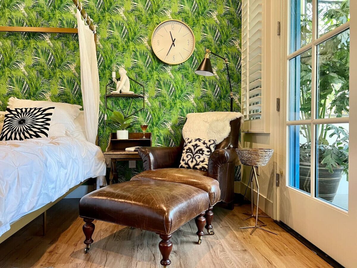 Um quarto com cama, uma cadeira, um relógio e uma janela com vista para plantas.
