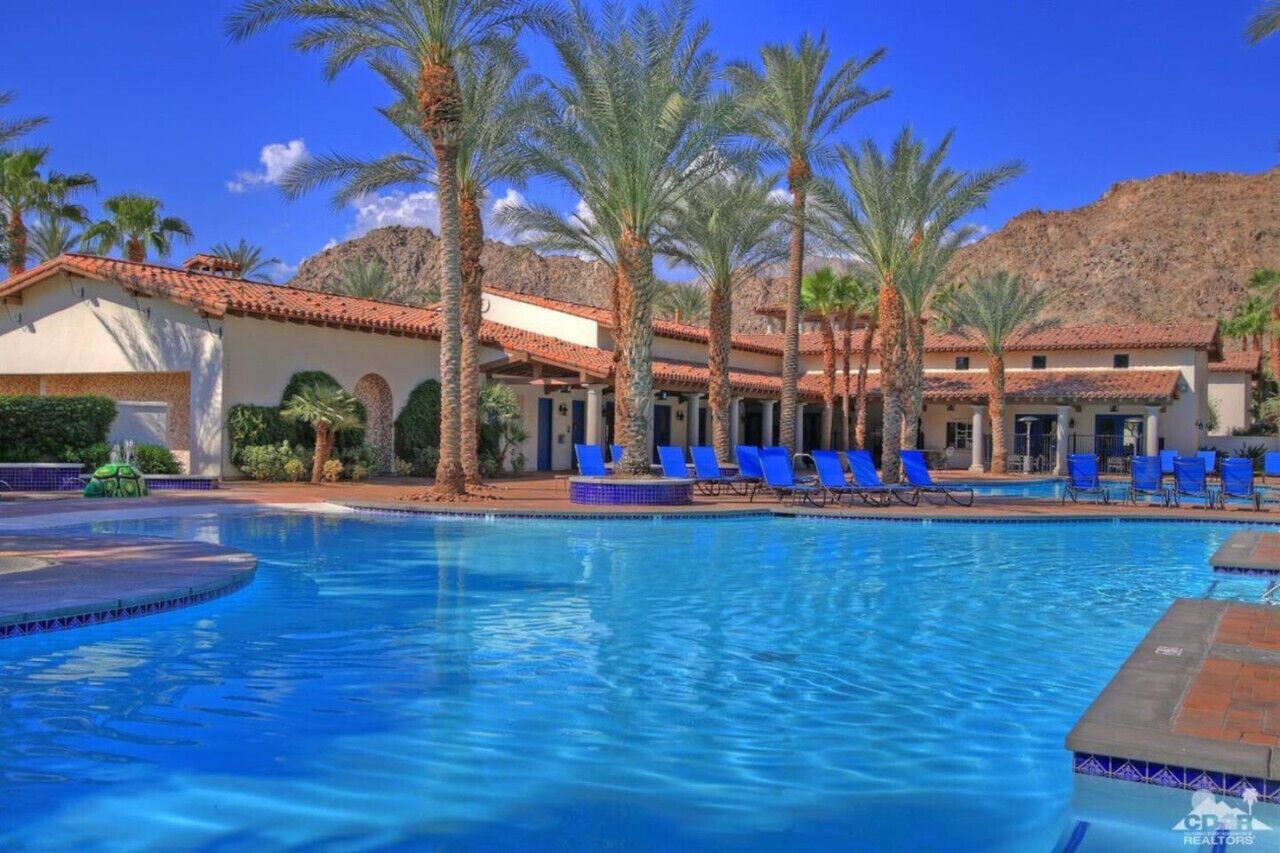 A swimming pool with blue water, surrounded by palm trees and a building with a tiled roof.