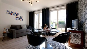 Ein modernes Wohnzimmer mit einer Couch, einem Esstisch mit Glasplatte und schwarzen Stühlen. Rechts befindet sich ein Fernseher auf einem Holzgestell.