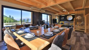 A spacious dining area with a long wooden table set for a meal, large windows offering a mountain view, and a cozy seating area with sofas and a television.