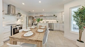 A modern kitchen with white cabinetry, stainless steel appliances, and a wooden dining table set for six. - North Shore Very Walkable to Ami fun (Anna Maria)