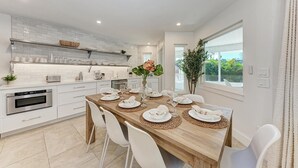 A modern kitchen with white cabinetry, a wooden dining table set for four, and a view of the outdoors through a large window. - North Shore Very Walkable to Ami fun (Anna Maria)