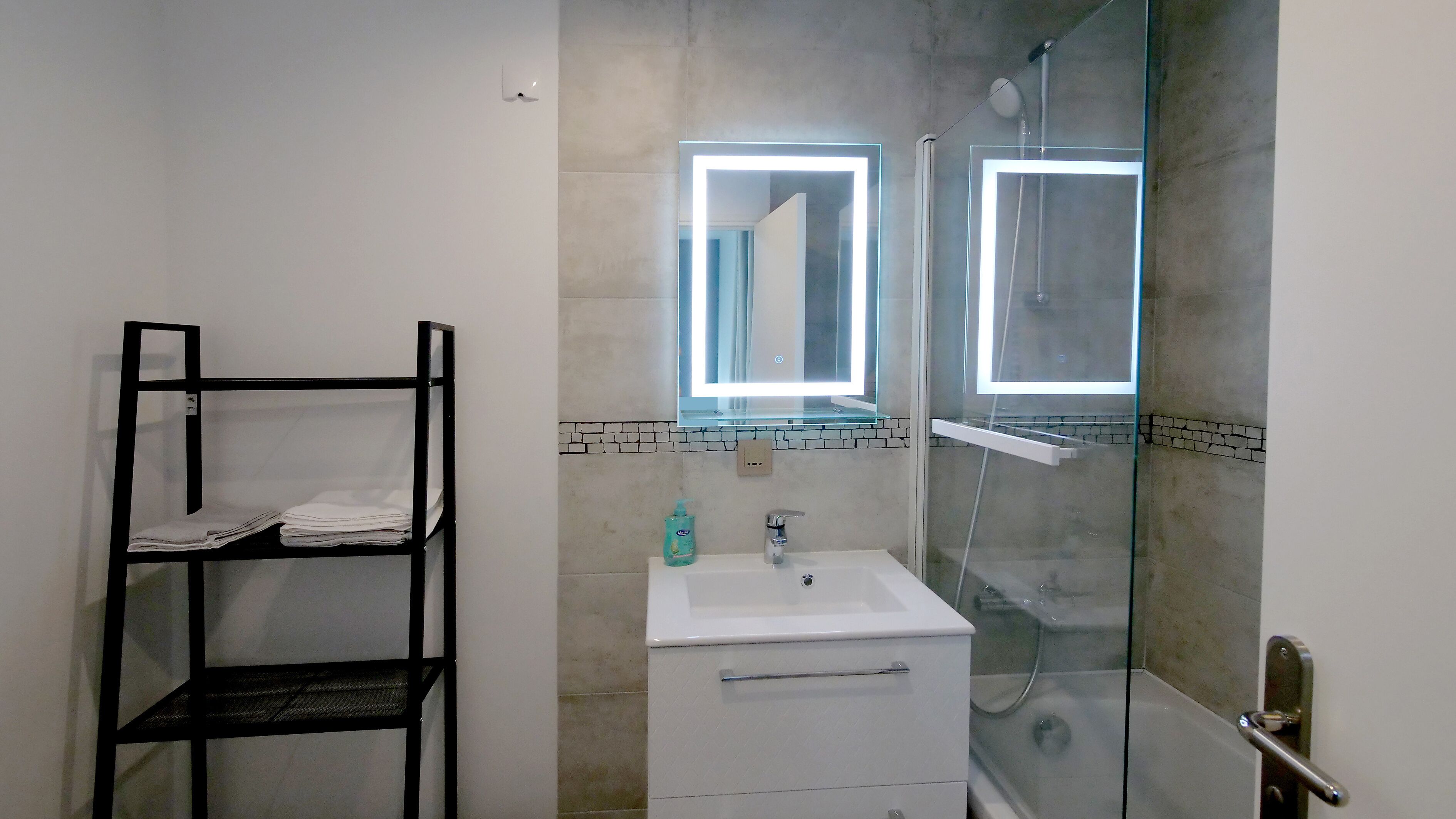 A bathroom with a white sink, a glass shower enclosure, and a black metal shelf unit.