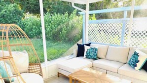 Une terrasse ensoleillée avec un fauteuil en osier, une table basse en bois et un canapé blanc agrémenté de coussins bleus.