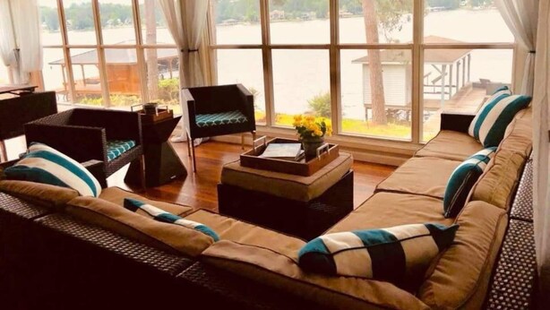 A spacious living room with a large sofa set, a small black chair, and a coffee table with a vase of flowers. Large windows provide a view of a lake and dock. - A Modern-Rustic Lake Retreat Get-Away! (Milledgeville)