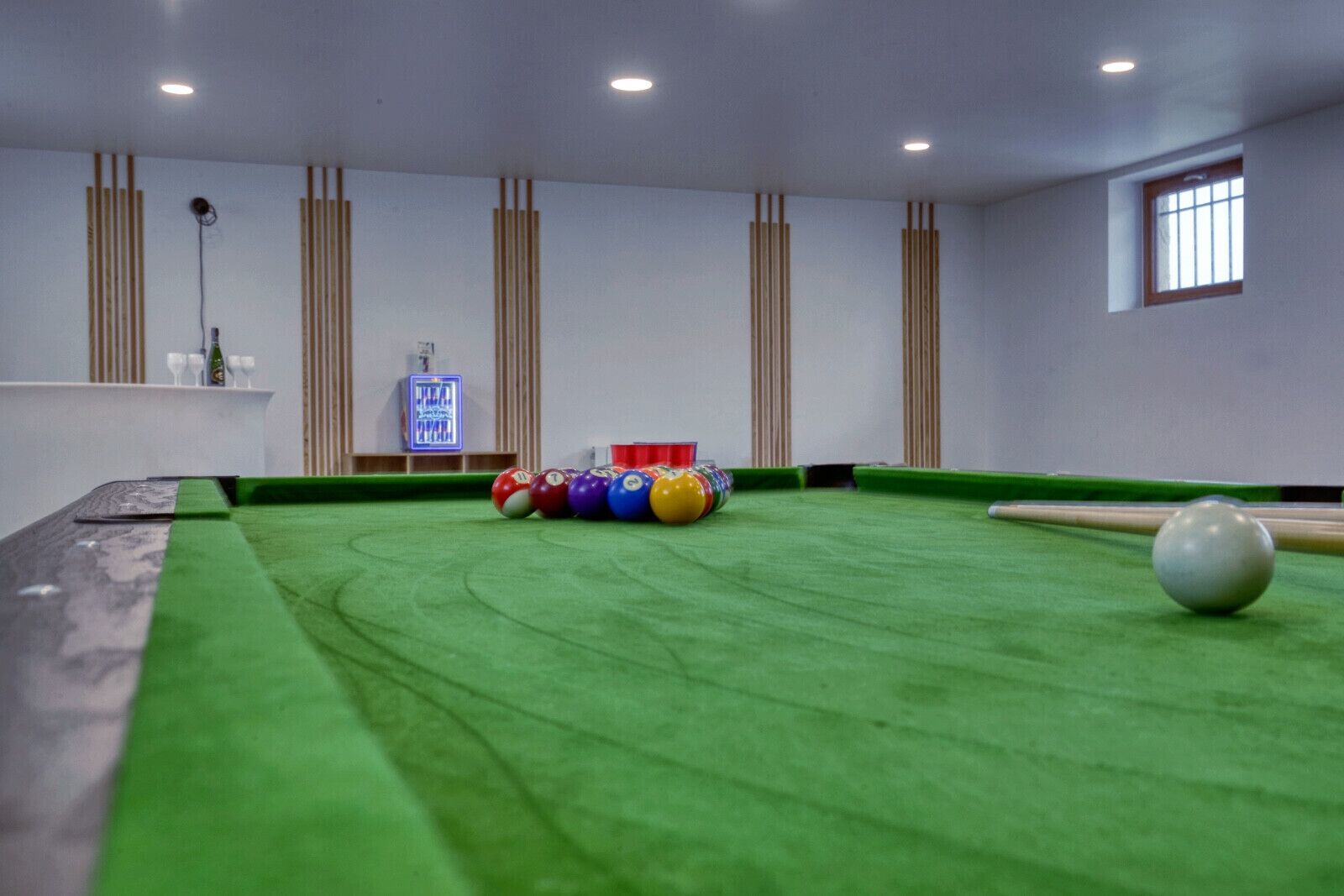 A pool table with balls and a cue ball, surrounded by beige walls and a window.